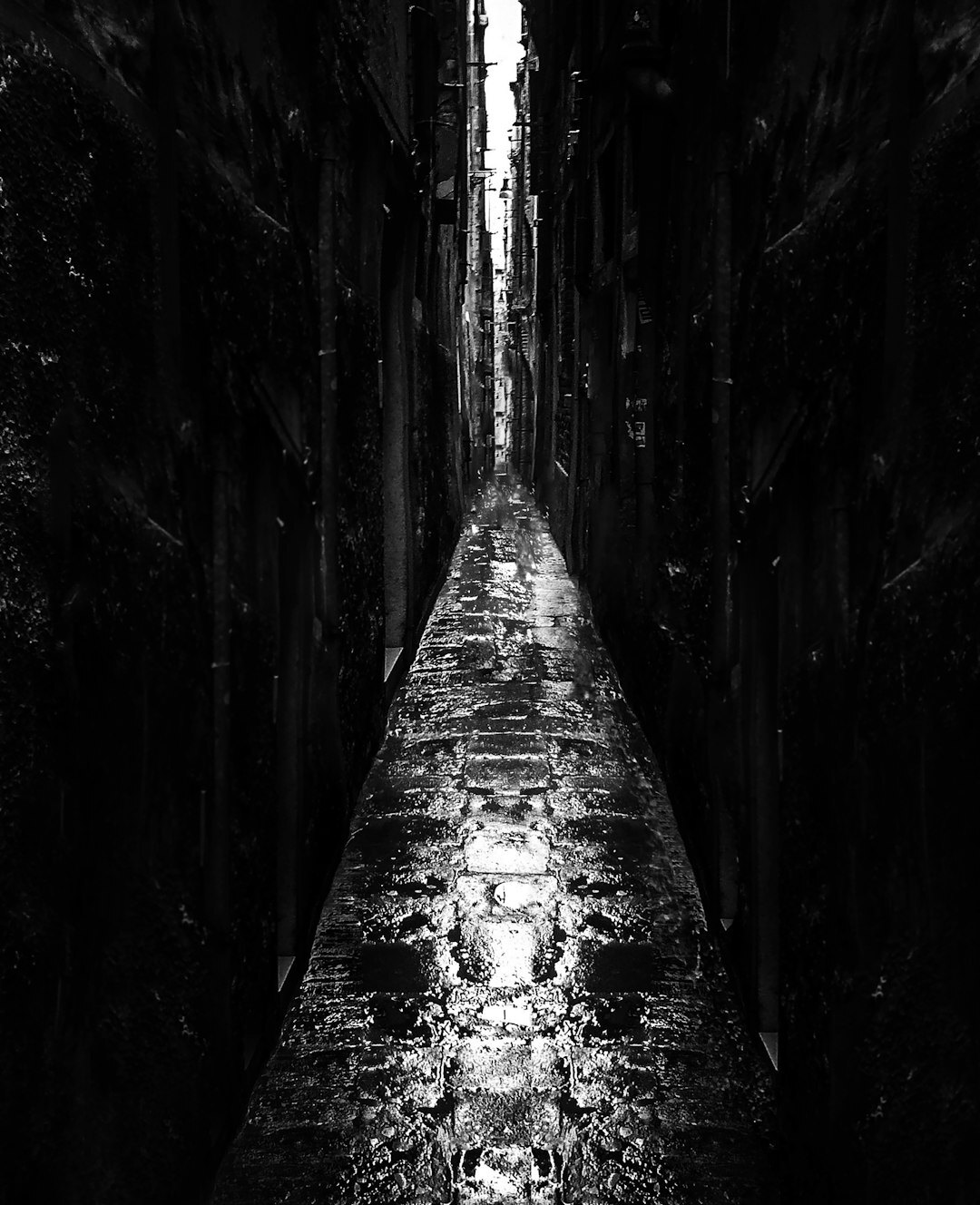 Dark narrow alley with wet pavement reflecting a single distant light