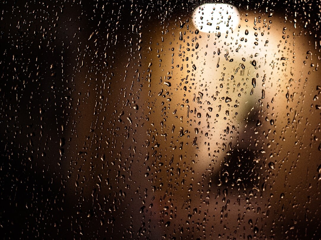 Rain droplets on a dark window with a warm reading lamp glowing in the background